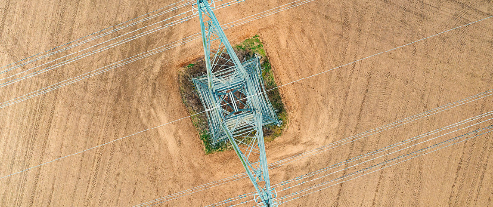 Aerial view of a high-voltage electricity pylon and power lines standing in the middle of a plowed agricultural field.